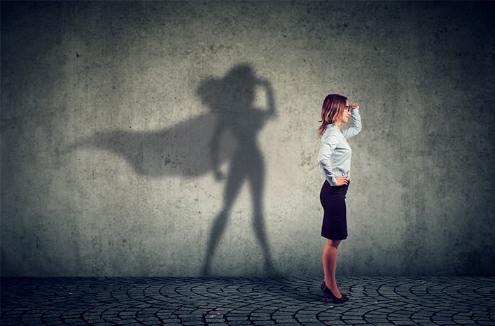 Femme en costume professionnel regardant au loin, ombre de super-héroïne en arrière-plan.